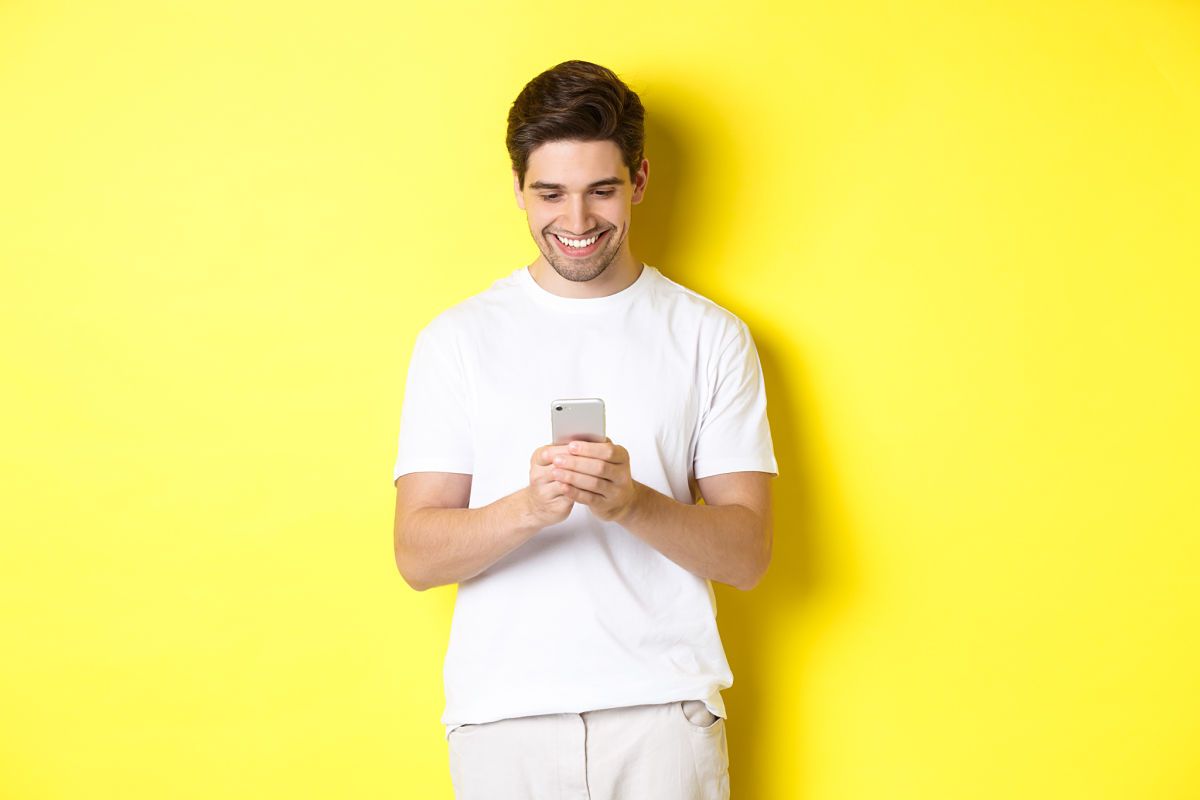 Young man reading text message on smartphone, looking at mobile phone screen and smiling, standing in white t-shirt against yellow background apps para aprender idiomas